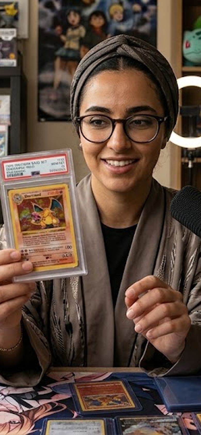 Dana holding a graded Charizard to camera at her desk in Manama, Arabic chat on the monitor beside her